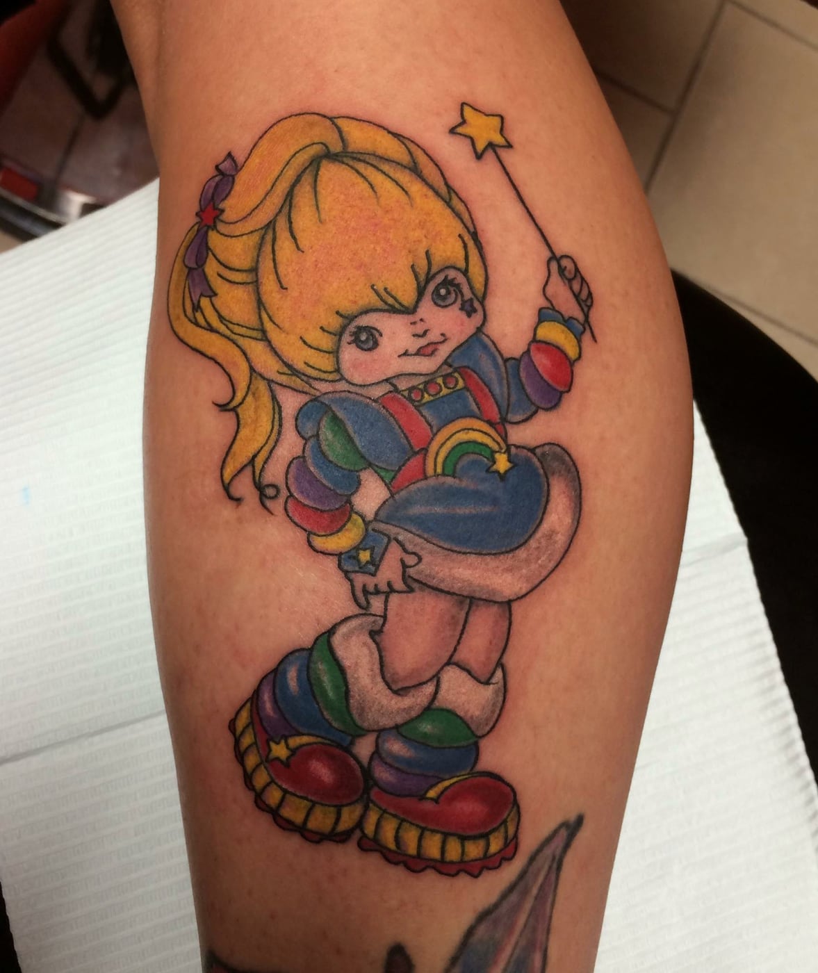 Bright new school color tattoo of a cartoon character in rainbow clothing holding a star wand, on a lower leg