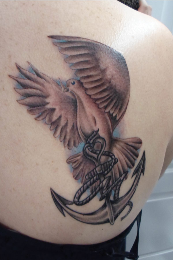 Black and grey dove and anchor tattoo on a shoulder blade, wings rendered in soft greywash over a pale blue background wash
