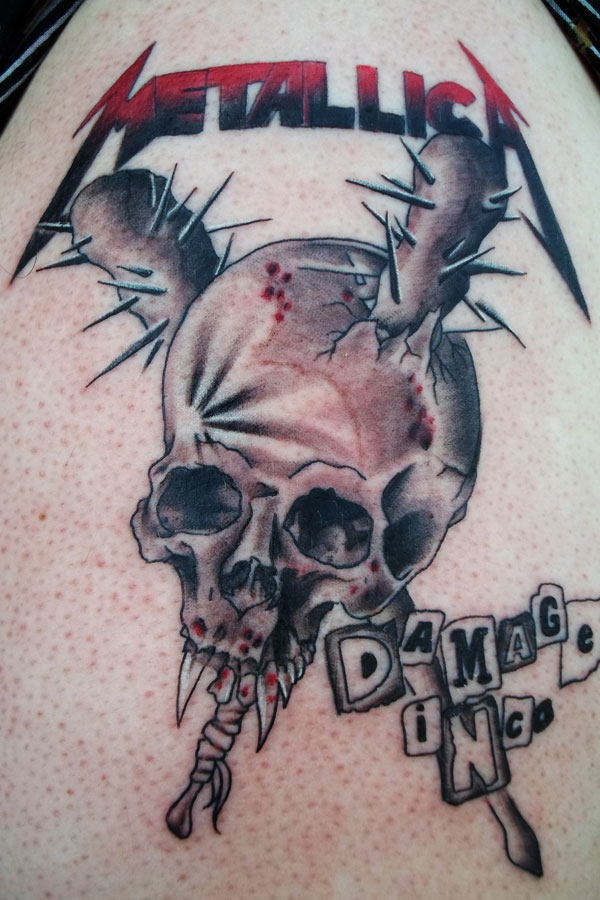 Realistic tattoo of a cracked skull pierced by spikes with lettering above and below it, rendered in grey with red accents