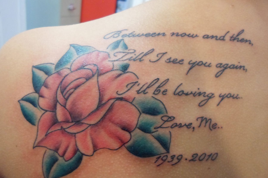 Memorial shoulder tattoo of a pink rose with teal leaves beside four lines of script and a pair of remembrance dates