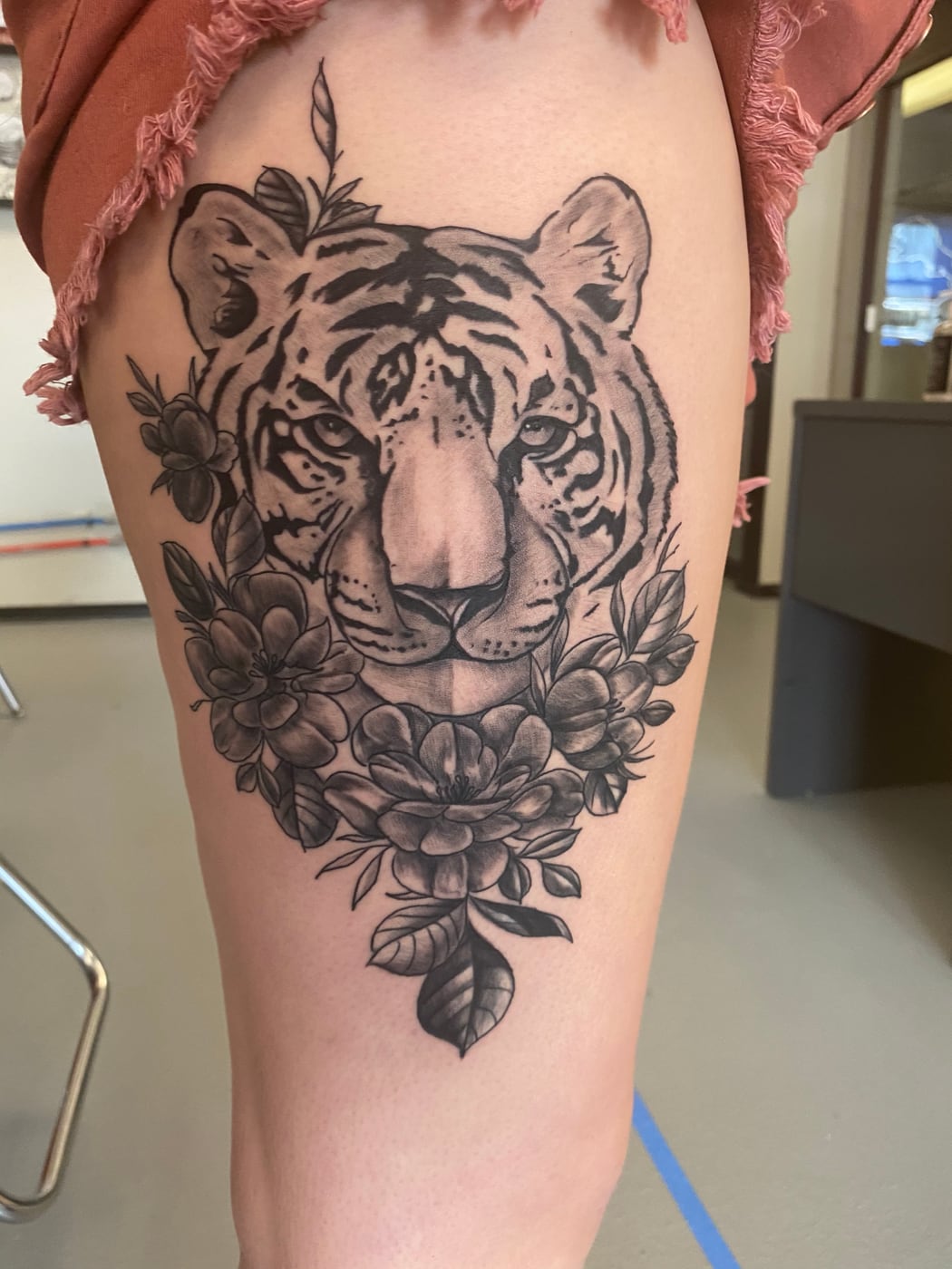 Realistic black and grey white tiger's head framed by peonies and foliage, covering the front of a thigh